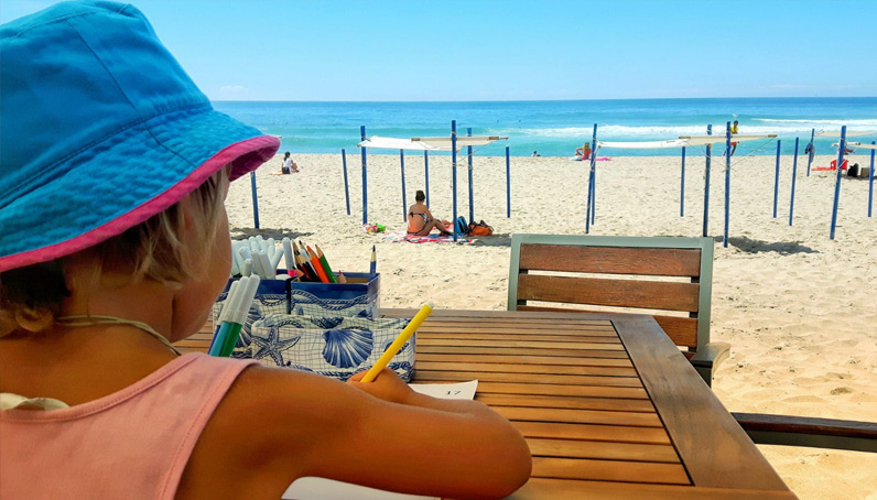 Ein Mädchen malt bei strahlendem Sonnenschein an einem Tisch am Strand.