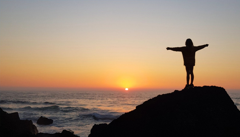 Ein Mädchen steht mit ausgebreiteten Armen auf einem Felsen an der Küste und umarmt den Sonnenuntergang während sie auf das Meer blickt.