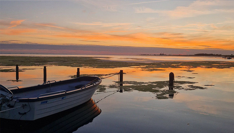 Ein kleines Motorboot liegt ruhig am Ufer einer Bucht über deren spiegelglatten Wasser die Sonne untergeht.